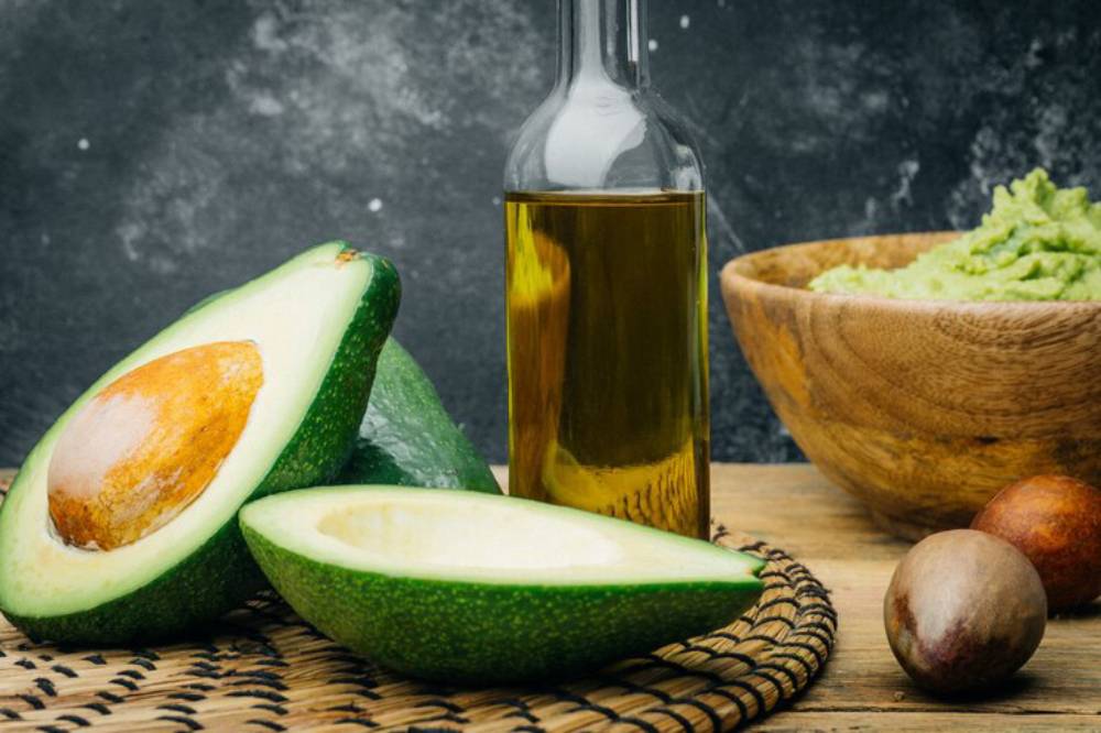 Half-cut avocados next to a bottle of avocado oil and a wooden bowl of guacamole, set on a woven mat with dark background.