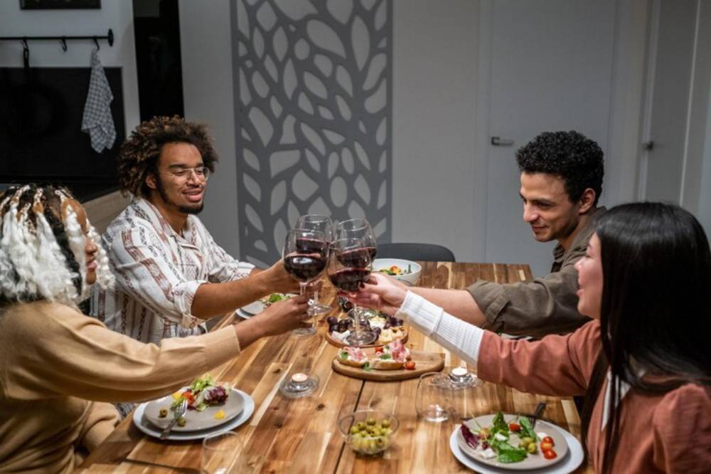 Three women prepare a colorful spread of appetizers and drinks on a rustic table, surrounded by plants and a warm interior setting.