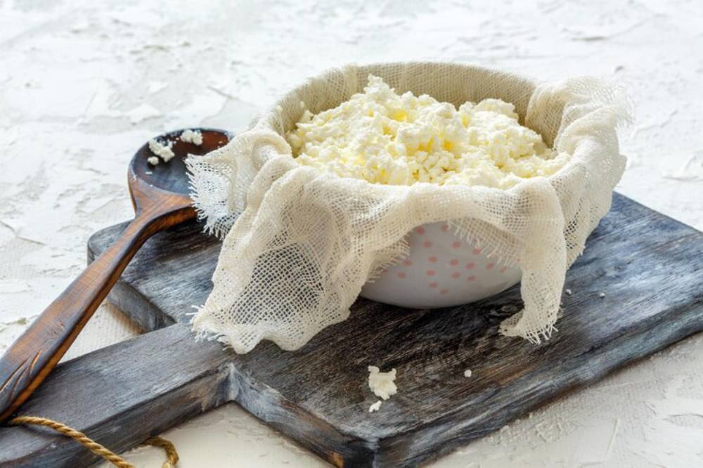 A bowl of freshly made cottage cheese draped with a cheesecloth on a wooden cutting board, accompanied by a wooden spoon.