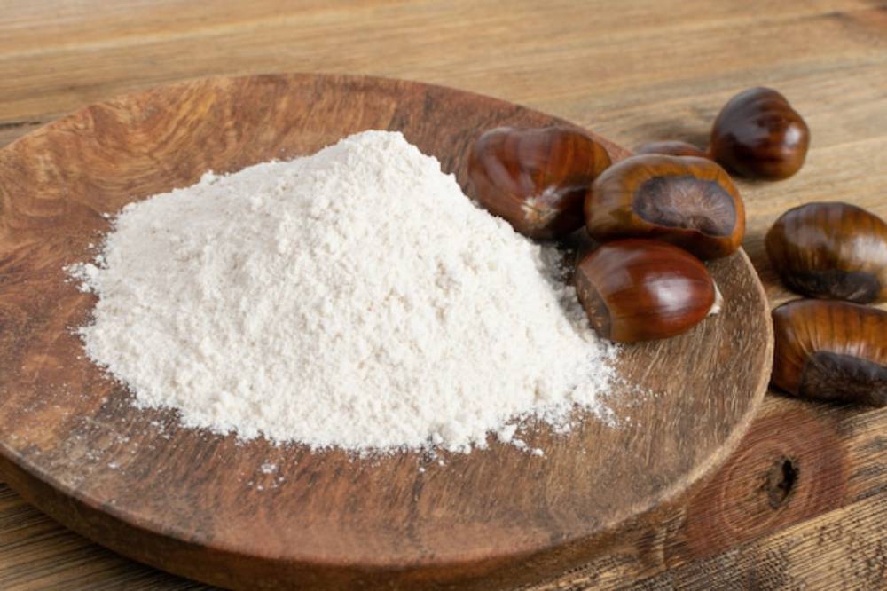A mound of chestnut flour rests on a wooden plate, surrounded by glossy, unpeeled chestnuts, set against a rustic wooden surface.
