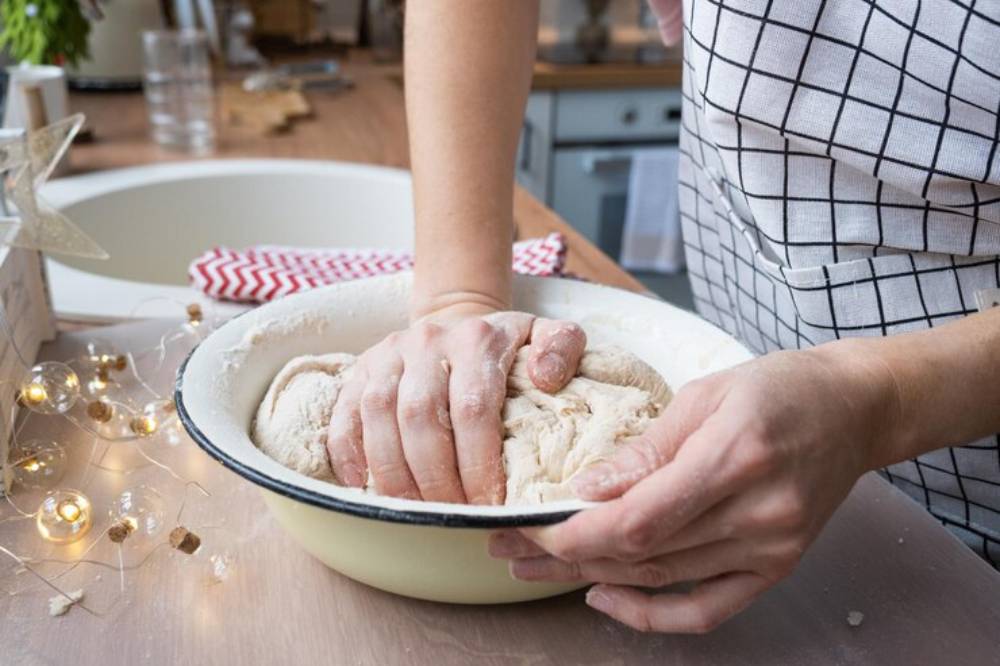 A person kneads dough in an enamel bowl, with flour-dusted hands, on a wooden countertop adorned with decorative lights.