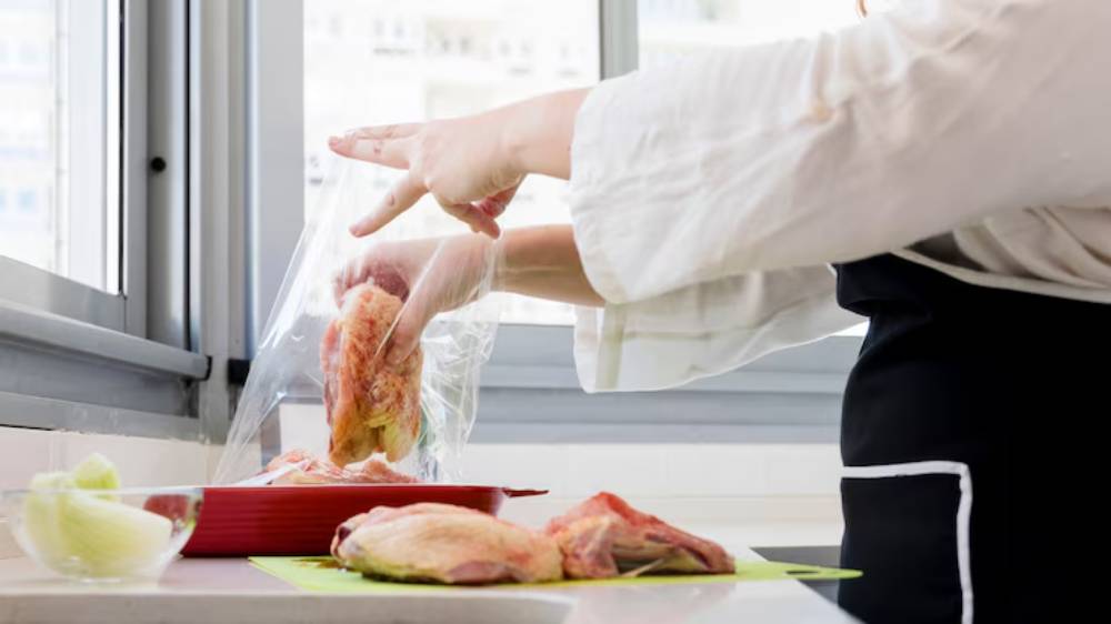 A person in an apron is placing raw chicken into a plastic bag while preparing ingredients in a bright kitchen.
