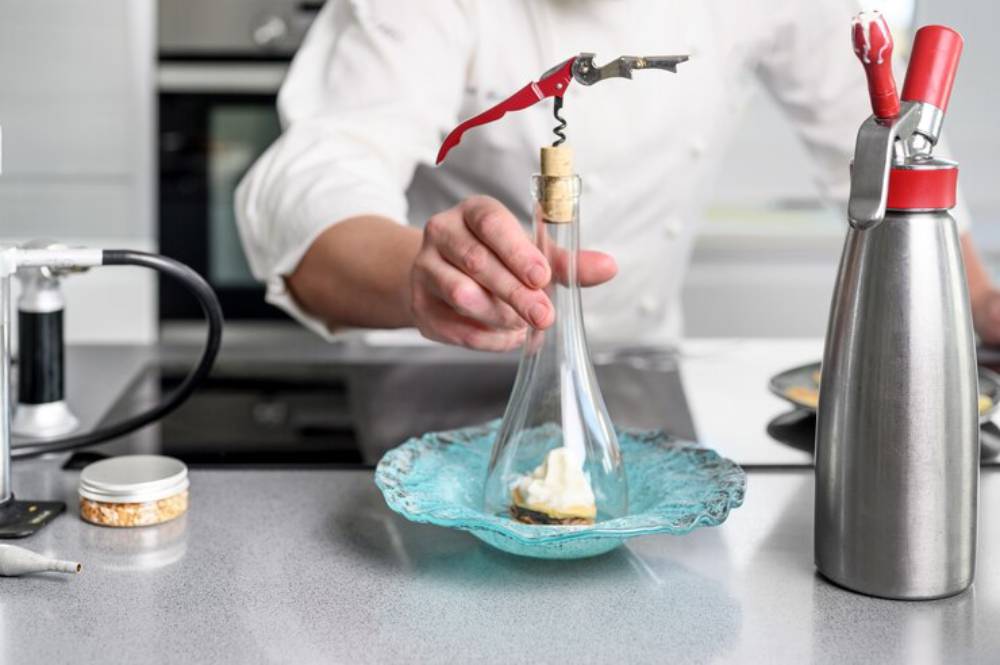 A chef uses a corkscrew to secure a cork in a glass flask, preparing a culinary dish with cream on an ornate blue plate.
