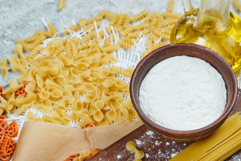 A bowl of flour surrounded by various pasta types and a bottle of olive oil, set on a textured surface.