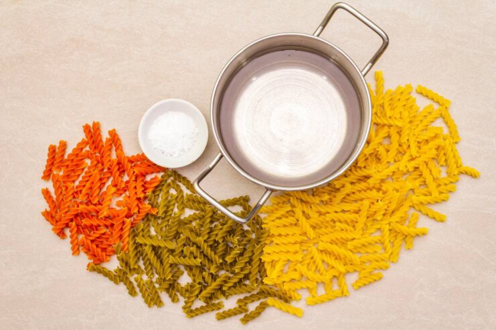 A stainless steel pot sits empty beside colorful spiraled pasta in orange, green, and yellow, alongside a small bowl of salt.