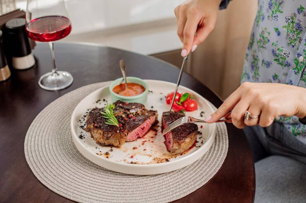 A person cuts into a perfectly cooked steak on a white plate, accompanied by cherry tomatoes and dipping sauce, with a glass of red drink nearby.