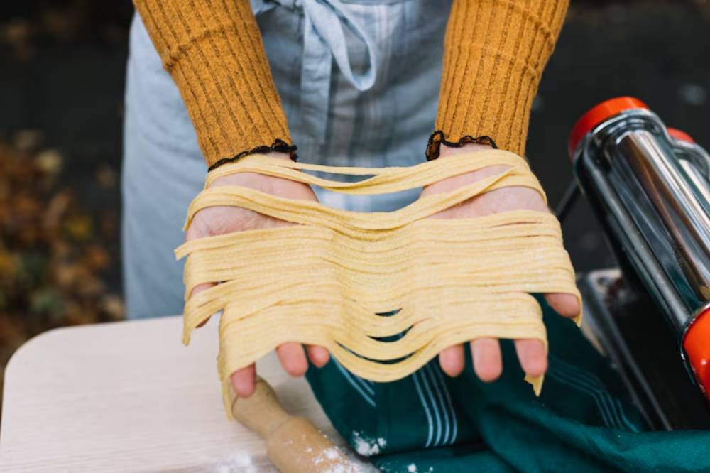 A person holds freshly made, long strands of pasta in their hands, showcasing the homemade dough in a cozy kitchen setting.
