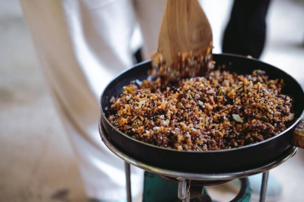A wooden spoon stirs a colorful mixture of grains and seeds in a frying pan on a stovetop, showcasing a vibrant cooking scene.