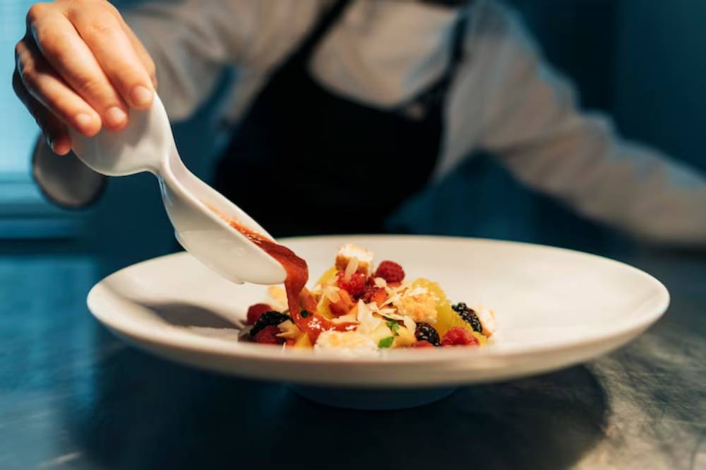 A person elegantly drizzles sauce over a vibrant dessert plated with berries and garnishes, showcasing culinary artistry.