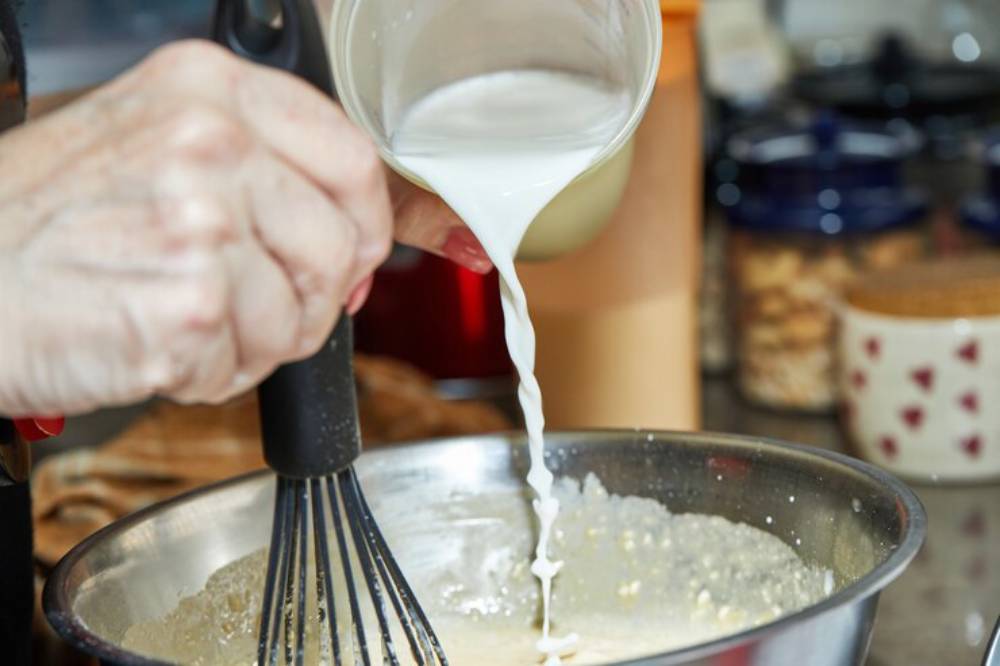 A hand pours milk from a glass into a mixing bowl containing batter, with a whisk nearby, in a cozy kitchen setting.