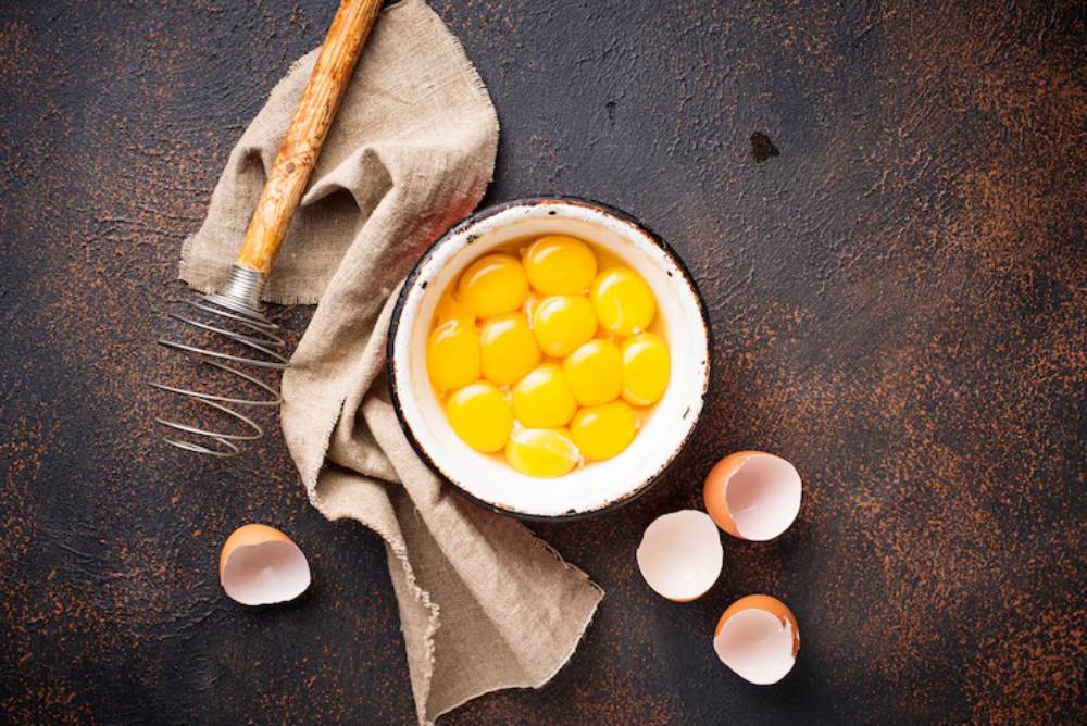 A bowl filled with fresh egg yolks surrounded by broken eggshells and a whisk on a dark textured surface.