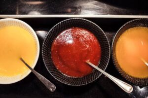 Three bowls of sauces: a creamy yellow sauce, a glossy red sauce, and a light brown sauce, each with a spoon for serving.