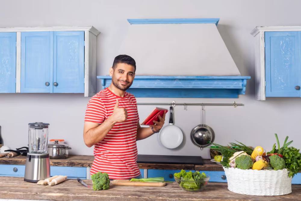 A person in a red-striped shirt stands in a bright kitchen, holding a tablet, surrounded by fresh vegetables and a blender on the counter.