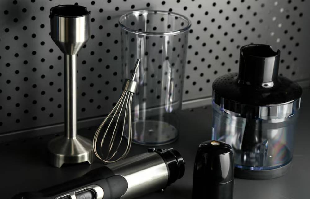 A collection of kitchen tools including a blender, whisk, and measuring cup, arranged on a pegboard background.
