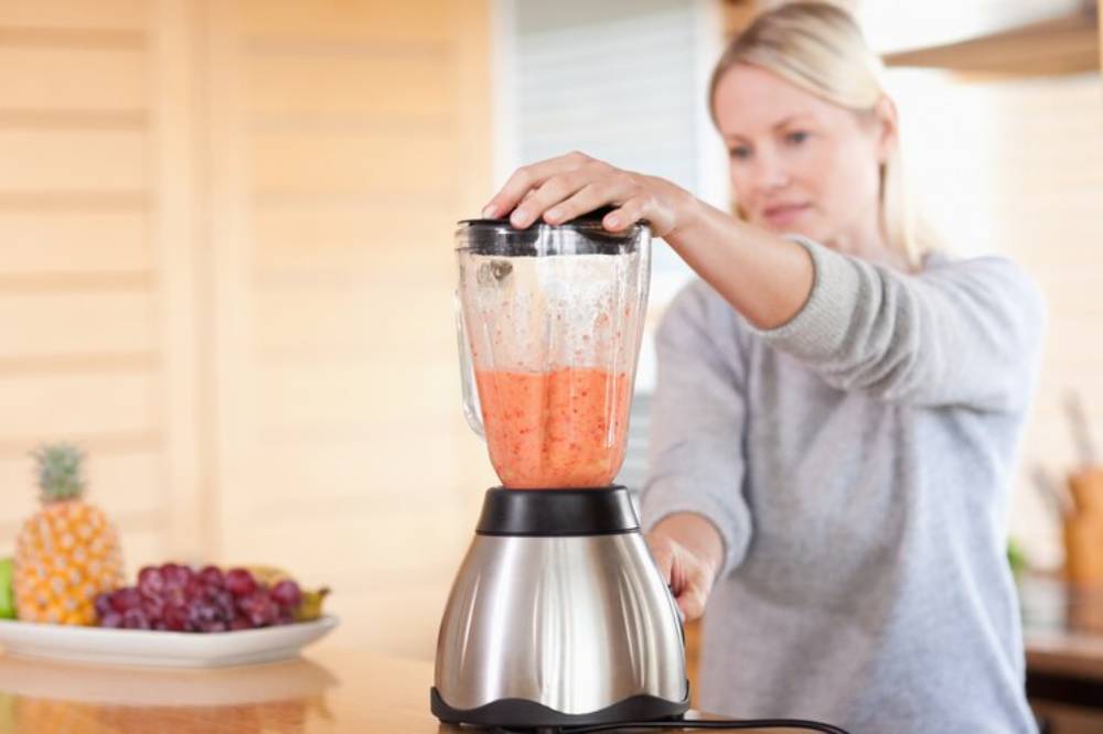 A person blends a bright orange mixture in a modern blender, surrounded by fresh fruits like grapes and pineapple on a wooden countertop.