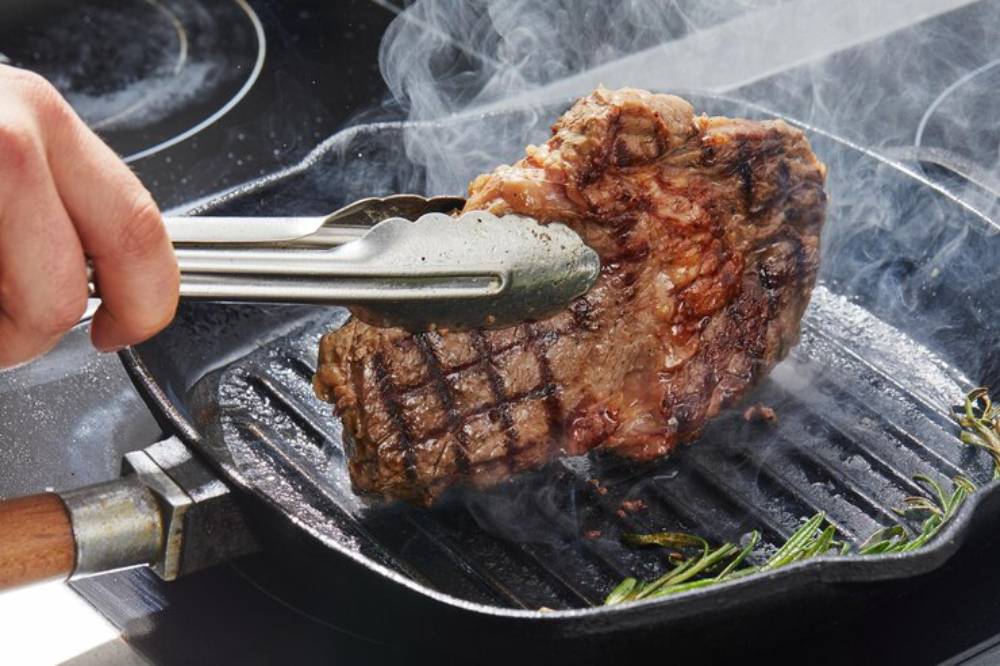 A hand using tongs to flip a sizzling steak on a grill pan, with steam rising and fresh herbs nearby.