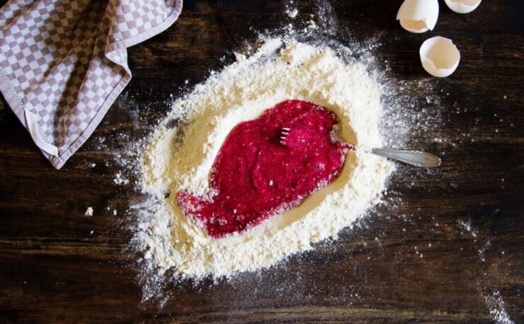 A mound of flour surrounds a vibrant red berry mixture, with a fork resting in the mixture, and eggshells nearby on a wooden surface.