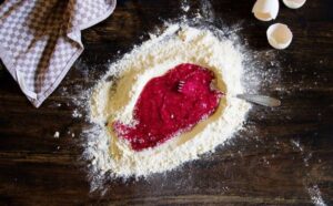 A mound of flour surrounds a vibrant red berry mixture, with a fork resting in the mixture, and eggshells nearby on a wooden surface.
