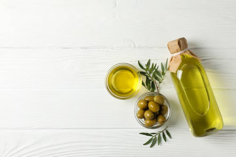 A bottle of olive oil alongside a glass with oil, a bowl of green olives, and olive leaves on a white wooden surface.