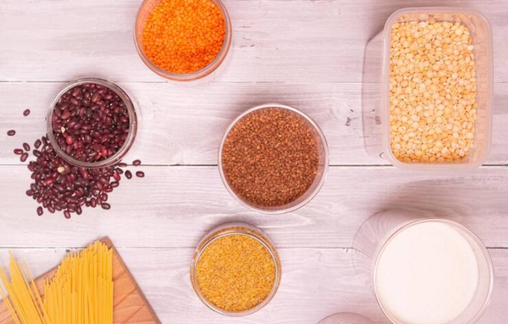Assorted grains and legumes in glass containers on a wooden surface, including red lentils, beans, and spaghetti.