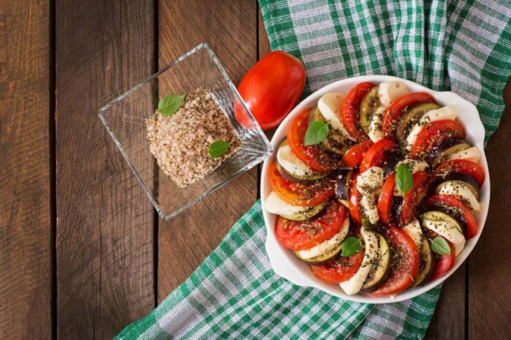 A colorful platter of tomato and eggplant slices garnished with herbs, beside a glass dish of seasoning and a fresh tomato on a checkered cloth.