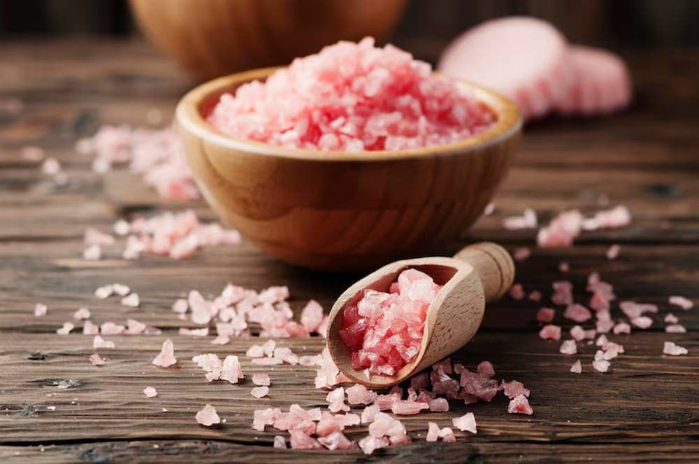 A wooden bowl filled with Himalayan pink salt, alongside a wooden scoop, scattered salt crystals on a rustic wooden surface.