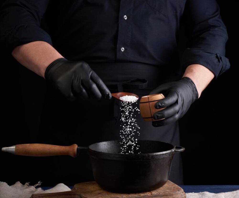 A chef in a black shirt and gloves pours salt from a wooden scoop into a cast iron pot on a dark background.
