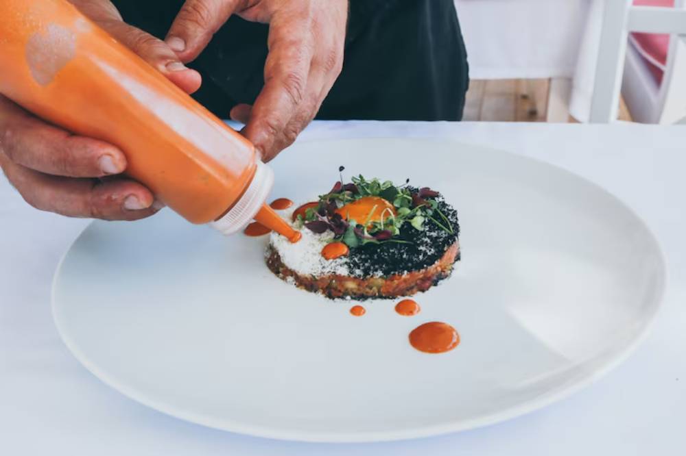 A chef decorates a gourmet dish with sauce, featuring a bright orange egg yolk nestled on a bed of greens and black ingredients.