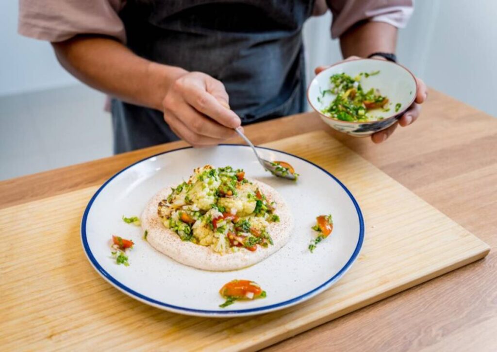 A chef meticulously garnishes a plate of cauliflower and vegetables atop a creamy base, adding finishing touches from a bowl.
