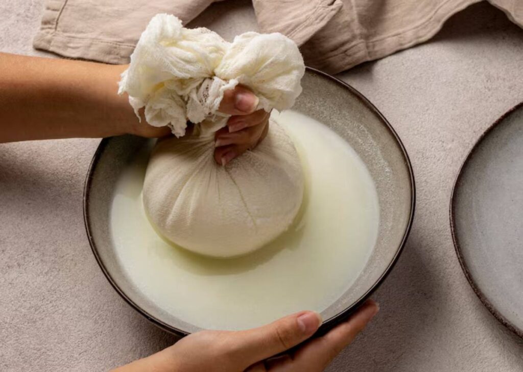 A person squeezes a cloth bundle containing pressed cheese over a bowl, extracting fresh whey. Light colors and soft textures are present.