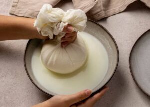 A person squeezes a cloth bundle containing pressed cheese over a bowl, extracting fresh whey. Light colors and soft textures are present.
