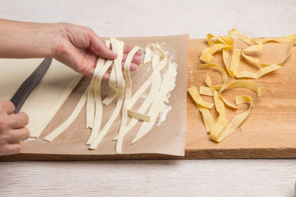 A hand holds freshly cut pasta strips over parchment paper, with more pasta resting on a wooden board beside it.