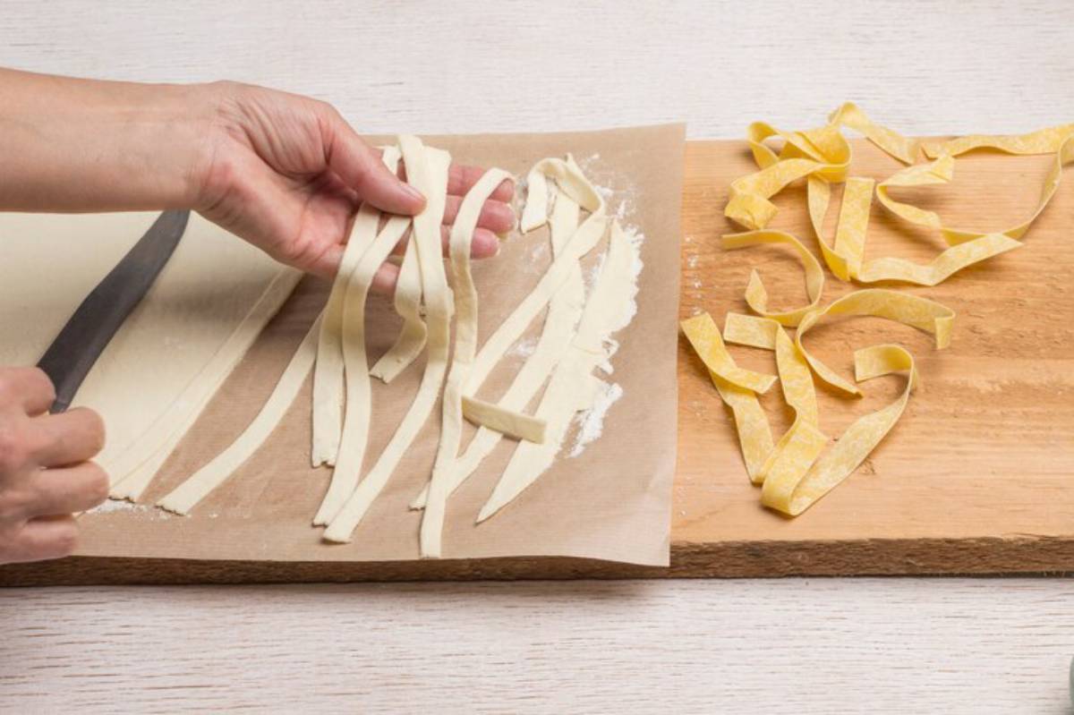 A hand holds freshly cut pasta strips over parchment paper, with more pasta resting on a wooden board beside it.
