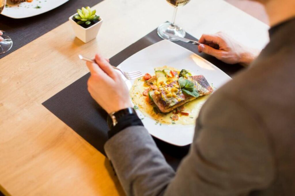 A person in a suit is enjoying a beautifully plated dish of fish with vegetables, garnished with herbs, and a glass of wine.