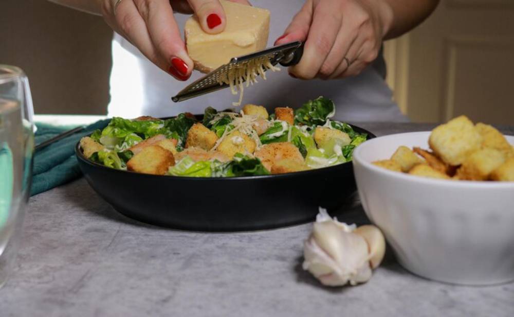 A person grating cheese over a fresh salad in a black bowl, with croutons and garlic nearby on a gray countertop.