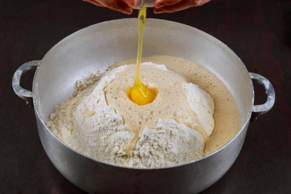 A hand cracks an egg into a mound of flour and batter in a large silver mixing bowl on a dark surface.