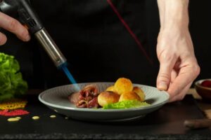 A chef using a culinary torch to caramelize meat on a plate with golden roasted potatoes and fresh lettuce in a dark setting.
