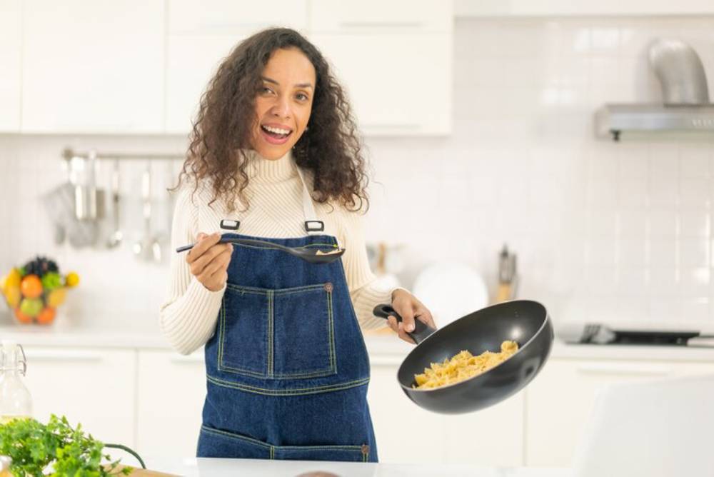 A person in a cozy kitchen, wearing a sweater and denim overalls, skillfully stirs pasta in a frying pan. Fresh ingredients nearby.