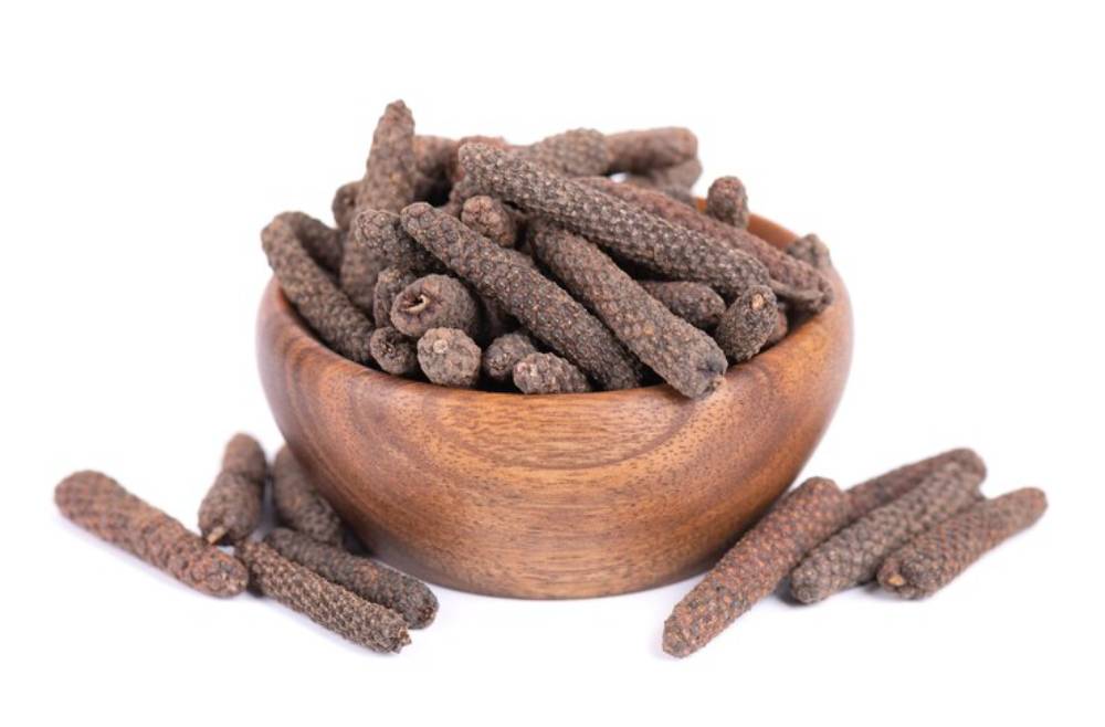 A wooden bowl overflowing with long, dark-brown spices, resembling sticks or pods, placed on a white background.