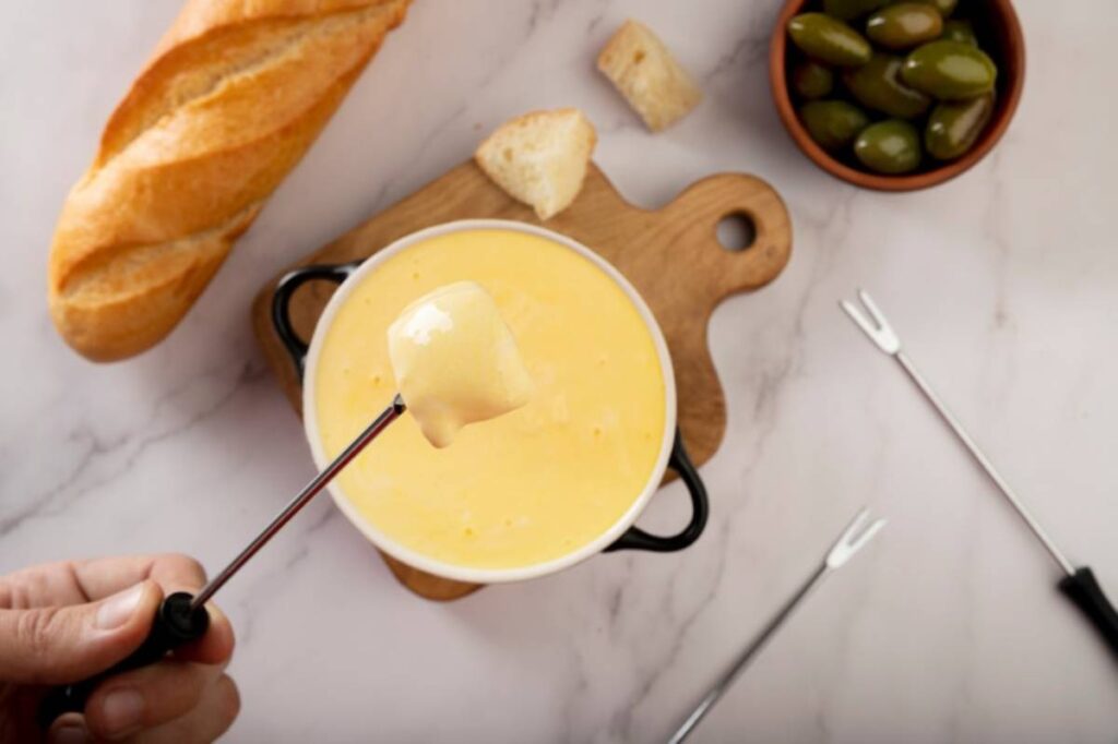 A hand holding a skewer with Béarnaise above a pot , accompanied by bread and olives on a wooden board.