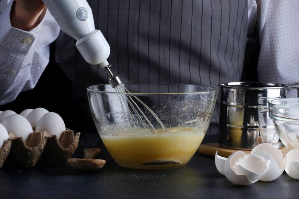 A person in a pinstripe shirt uses a hand mixer on a bowl of sauce, with eggs and broken shells nearby.