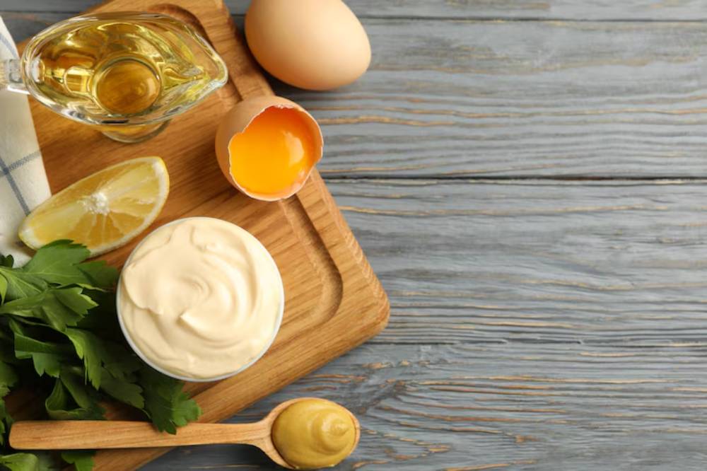 Ingredients arranged on a wooden board: olive oil, lemon, egg, mustard, mayonnaise, and fresh parsley.