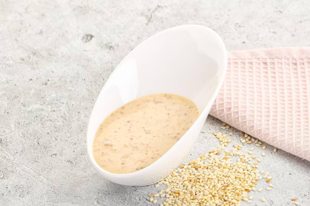 A white bowl holds sesame butter, surrounded by scattered sesame seeds and a pink kitchen towel on a stone surface.