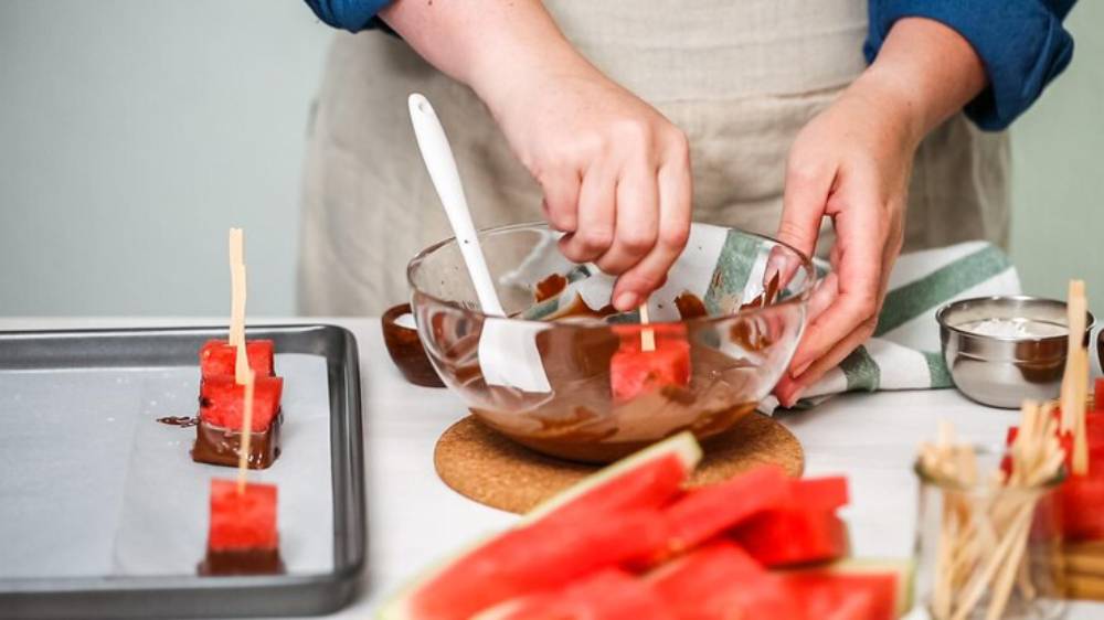 A person coats watermelon slices with melted chocolate using a spatula, surrounded by fresh watermelon and preparation tools.