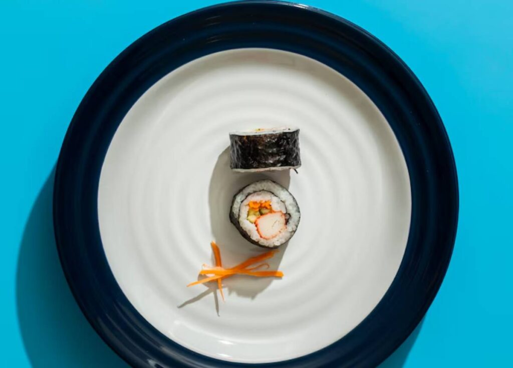 A close-up of a sushi roll on a white plate with a blue rim, accompanied by thin orange carrot garnish.