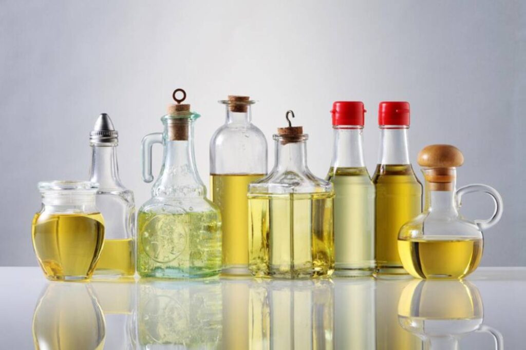 Various bottles of oil, including glass and plastic, with different shapes and caps, arranged on a reflective surface.