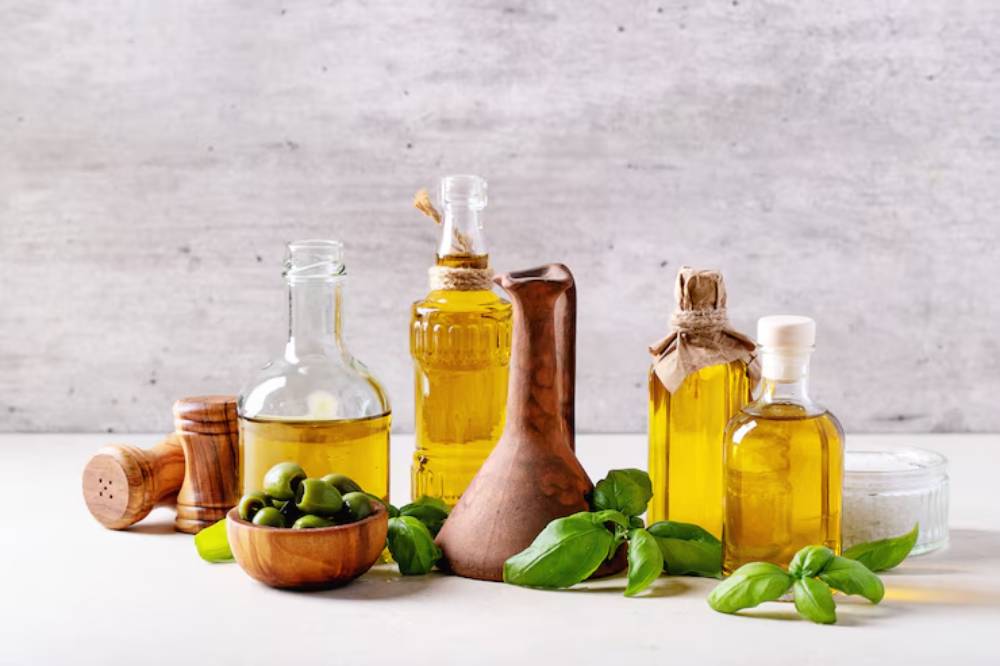 A collection of olive oils in glass and clay bottles, green olives in a wooden bowl, and fresh basil leaves on a light background.