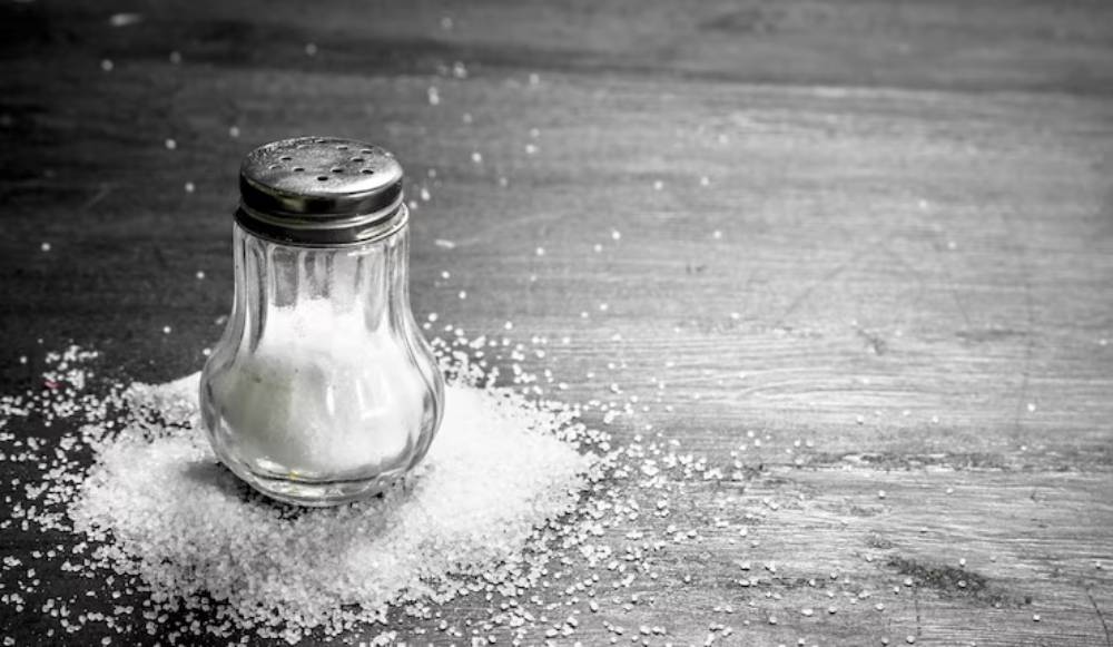 A glass salt shaker sits atop a scattered mound of salt on a wooden surface, captured in a black and white image.