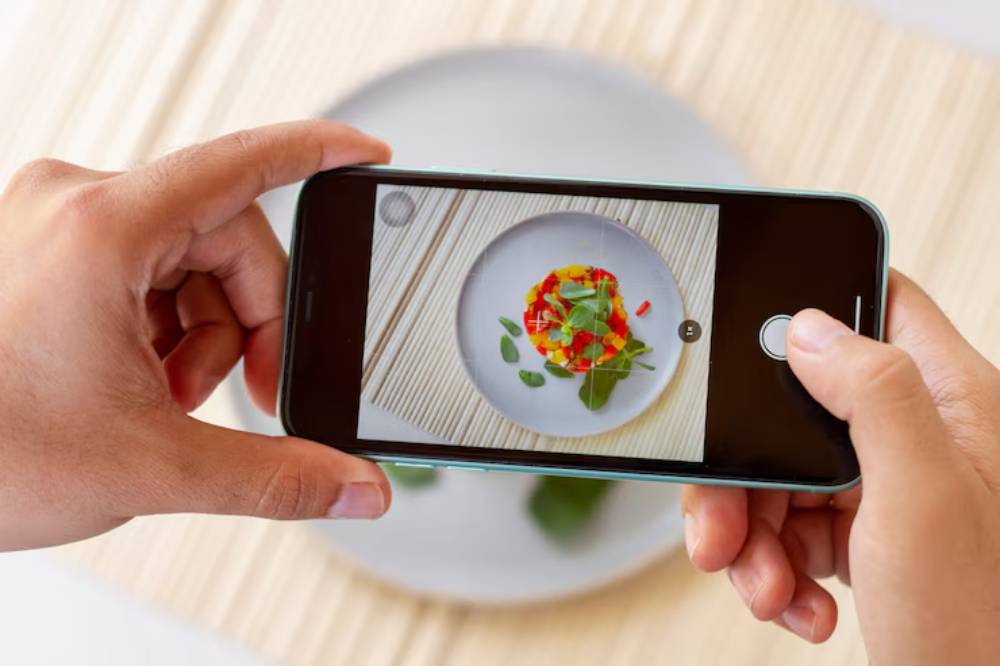 A person holds a smartphone capturing an artistic plate of colorful food garnished with greens.