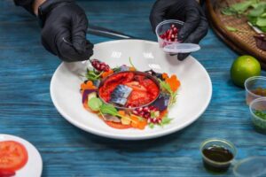 A chef in black gloves artfully arranges a colorful salad with pomegranate, herbs, and vegetables on a white plate.